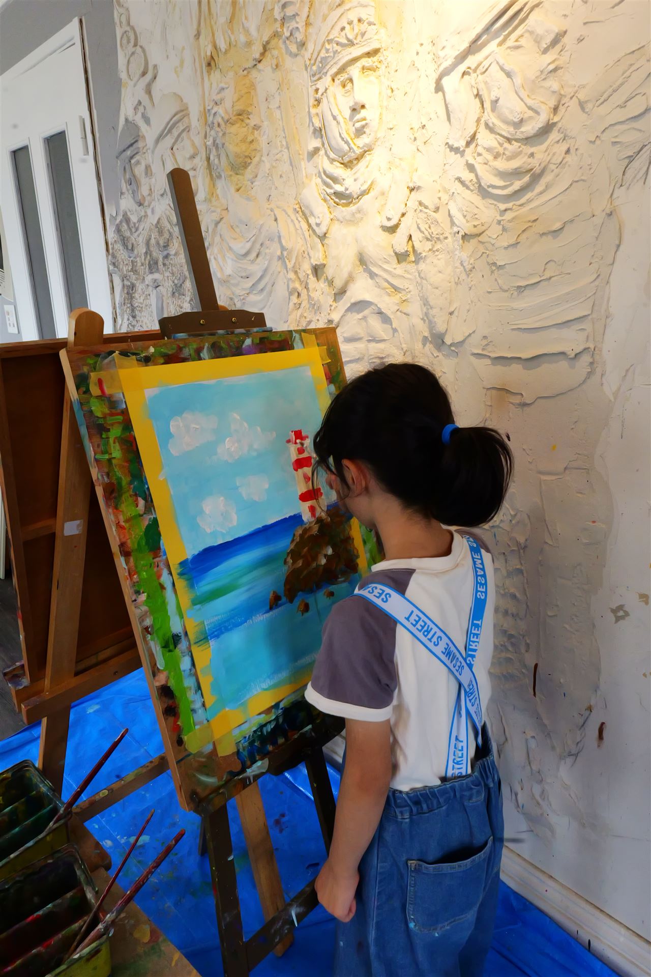 レリーフの壁の前でイーゼルに向かい灯台の絵を描く子ども